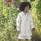 A child beams with joy in a natural garden setting, wearing Save The Duck's lightweight, cream-colored rain jacket. The jacket's eco-friendly design is perfect for playful adventures, providing both comfort and protection. The backdrop of lush greenery and vibrant pink flowers captures the essence of spring, aligning with the brand's vision of harmony with nature. | Save The Duck | Animal Free Elegant Duvets for Men and Women