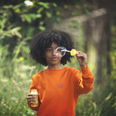 Curious child in a vibrant orange, animal-free fabric sweatshirt blowing a bubble, surrounded by greenery | Save The Duck | Animal Free Elegant Duvets for Men and Women
