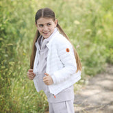 A young girl smiles brightly in a verdant outdoor setting, clad in a pristine white Save The Duck puffer jacket. The jacket's signature orange logo adds a playful touch to the clean design, symbolizing the brand's cruelty-free commitment. Her coordinated light grey hoodie and pants suggest a comfortable yet stylish ensemble, suitable for the refreshing outdoors of spring. | Save The Duck | Animal Free Elegant Duvets for Men and Women