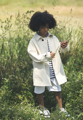 Curious child exploring nature while dressed in a chic beige SaveTheDuck trench coat over a striped shirt and white shorts, complemented by classic white sneakers. | Save The Duck | Animal Free Elegant Duvets for Men and Women