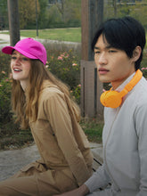 Young woman in a stylish beige SaveTheDuck jacket and pink cap laughing, accompanied by a young man in a sleek white jacket with vibrant orange headphones around his neck, representing cruelty-free fashion. | Save The Duck | Animal Free Elegant Duvets for Men and Women