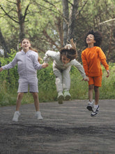Four children in playful harmony, jumping and holding hands in a lush park setting. Two boys, one in a dark navy hoodie and shorts, the other in an orange sweatshirt and shorts, leap with joy. Between them, a girl in a light grey tracksuit shares their excitement. On the far right, another girl in a cream jacket and pants takes flight with glee. Their casual, eco-friendly outfits are ideal for outdoor fun, embodying the spirit of carefree childhood. | Save The Duck