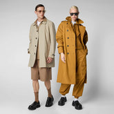 Two models present a sleek, modern take on eco-friendly outerwear from Save The Duck's Spring/Summer 2024 collection. On the left, a male model in a classic beige trench coat paired with a light brown ensemble exudes timeless style, while his counterpart on the right makes a bold statement in a long, mustard-colored raincoat over a matching set. Both complement their outfits with chunky black footwear and sunglasses, merging traditional and contemporary fashion elements. | Save The Duck | Animal Free Elegant Duvets for Men and Women
