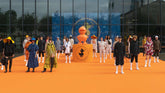 Models showcasing a diverse collection of animal-friendly jackets on a vibrant orange runway for Save The Duck, featuring a large duck mascot in the center. - Shop by look | Save The Duck