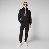 A man wearing a black hoodie with white drawstrings and matching black joggers from SaveTheDuck, standing in a studio setting. | Save The Duck | Animal Free Elegant Duvets for Men and Women
