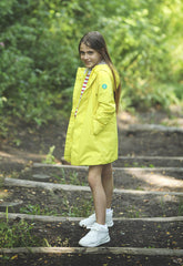 Smiling young girl in a bright yellow SaveTheDuck raincoat with a colorful striped lining, taking a playful stance on forest stairs, perfectly blending style with nature-friendly apparel. | Save The Duck | Animal Free Elegant Duvets for Men and Women