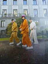Three models stride confidently in the rain, sporting Save The Duck's eco-friendly raincoats from the Spring/Summer 2024 collection. From left to right: a model in a mustard yellow coat, the center one draped in vibrant orange with a bright pink bucket hat, and the third in a classic white, embodying the brand's commitment to bold style and sustainability. Their coordinated white sneakers emphasize the collection's youthful and urban appeal, perfect for the conscious consumer on the go. | Save The Duck | Animal Free Elegant Duvets for Men and Women