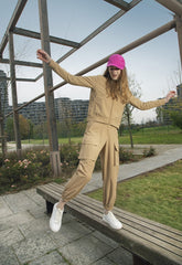 Fun and fashionable SaveTheDuck athleisure, featuring a woman in a beige tracksuit and striking pink cap, playfully balancing on a park bench with a backdrop of urban greenery. | Save The Duck