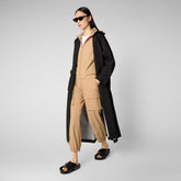 Model in SaveTheDuck's eco-friendly black overcoat and beige athleisure outfit, paired with white hoodie and black slides, striking a pose against a minimalistic grey background for a sleek, sustainable sportswear look. | Save The Duck