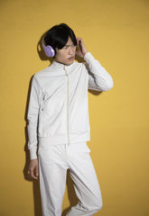 Man in a monochrome white SaveTheDuck tracksuit posing against a mustard yellow wall, complementing his outfit with lilac headphones for a striking contrast and a trendy athleisure vibe. | Save The Duck