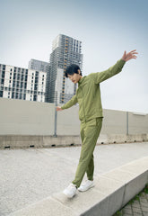 Dynamic SaveTheDuck athleisure style with a man in an olive tracksuit balancing on a city curb, blending eco-conscious fashion with urban exploration. | Save The Duck