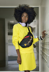 Vibrant SaveTheDuck athleisure with a young woman sporting a bright yellow sweatshirt and pleated skirt, accessorized with white headphones and a colorful waist bag, engaging with school lockers for an energetic, youthful look | Save The Duck