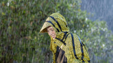 A woman cherishes a moment in the rain, wearing a vibrant animal-print, water-resistant jacket with a practical hood. The jacket's vivid yellow and black pattern stands out against the soft-focus greenery in the background. She is smiling gently, her face partially turned away, as raindrops cascade around her, highlighting the jacket's suitability for wet weather. Her demeanor suggests comfort and contentment in an eco-friendly attire that's both functional and fashionable. | Save The Duck