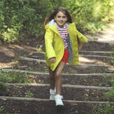 Happy young girl in a vibrant yellow eco-friendly jacket playfully descending outdoor stairs in a lush park | Save The Duck | Animal Free Elegant Duvets for Men and Women